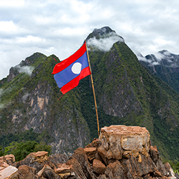 image du drapeau du Laos avec des couleurs rouge bleu et banche, planté au sommet d'une falaise