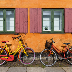 image de 2 vélos : un jaune et un rouge posé sur un mur de maison orange avec 2 volets en bois